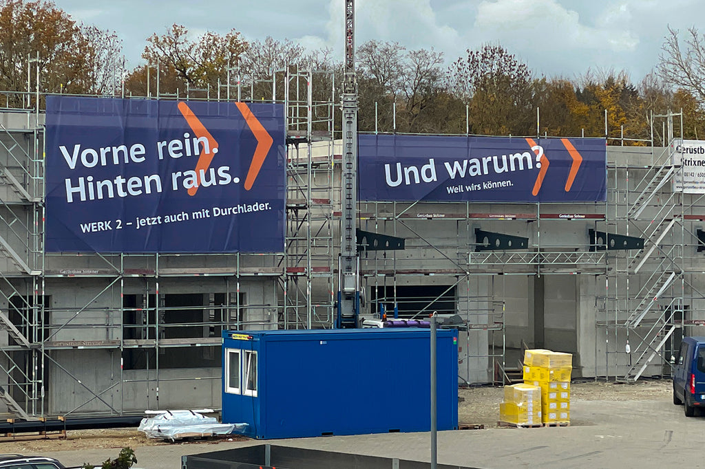 Zwei großformatige Meshbanner an der Fassade des neuen Werks von Butz Aufzüge. Die Banner mit dem Text ‚Vorne rein. Hinten raus.‘ und ‚Und warum? Weil wir’s können.‘ wurden von VIS24produziert.