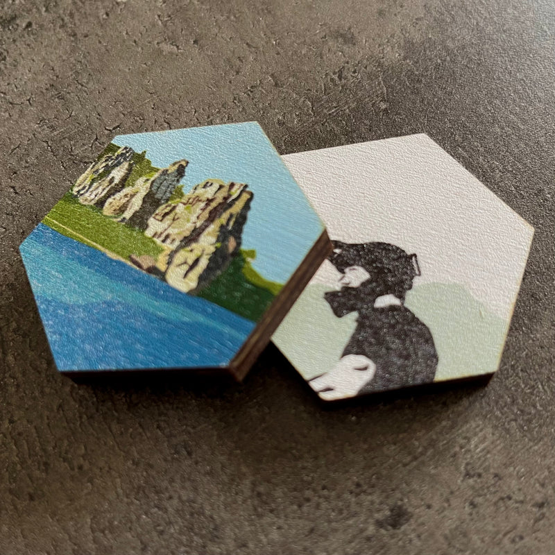 Zwei sechseckige Holzplatten mit farbigem Foto-Druck, eine mit einer Landschaftsszene und eine mit einer stilisierten Illustration einer Person mit Hut, auf einer dunklen Steinoberfläche liegend.