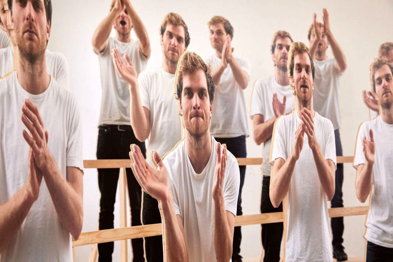 Mehrere identische Männer in weißen T-Shirts erscheinen in einer surrealen Szene, als ob sie klatschen und sich in einem Spiegelraum vervielfältigen.