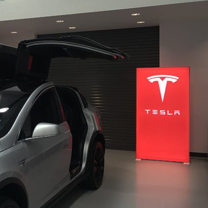 Beleuchteter Pixlip Go Rahmen mit Tesla-Logo im Autohaus, platziert neben einem Fahrzeug mit geöffneten Flügeltüren.